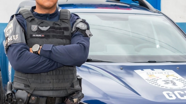 Agente da GCM de Carapicuíba posa com os braços cruzados em frente a uma viatura azul da corporação. Ele veste uniforme completo, colete tático e boina preta. Ao fundo, o muro da base da GCM, com portas e janelas. Foto: Prefeitura de Carapicuíba (Redes Sociais/Reprodução0