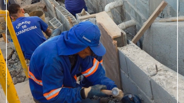#ParaQueTodosPossamVer: Servidores da Prefeitura de Carapicuíba realizam obra de zeladoria em uma viela. A imagem mostra trabalhadores uniformizados construindo uma escada de alvenaria entre muros residenciais. Um dos servidores, em primeiro plano, manuseia uma marreta sobre uma base de concreto. Ao fundo, outros servidores alinham blocos de cimento e preparam o local para continuidade da construção. A ação visa melhorar o acesso e a segurança dos moradores da região. Foto: Prefeitura De Carapicuíba (Redes Sociais/Reprodução)