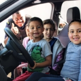 #ParaQueTodosPossamVer: Na imagem, um grupo de crianças está sentado dentro de um veículo, com um menino ao volante e as demais sorrindo ao redor. Ao lado de fora, um guarda civil uniformizado sorri e faz sinal de positivo com a mão. Foto: Prefeitura De Carapicuíba (Redes Sociais/Reprodução)