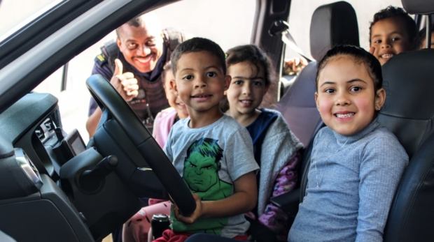#ParaQueTodosPossamVer: Na imagem, um grupo de crianças está sentado dentro de um veículo, com um menino ao volante e as demais sorrindo ao redor. Ao lado de fora, um guarda civil uniformizado sorri e faz sinal de positivo com a mão. Foto: Prefeitura De Carapicuíba (Redes Sociais/Reprodução)