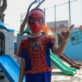 #ParaQueTodosPossamVer A imagem mostra um menino vestido de Homem-Aranha em um parquinho. Ele usa máscara e fantasia vermelha e azul, e faz o gesto clássico do herói com a mão, como se estivesse lançando uma teia. Ao fundo, há escorregadores coloridos e um muro pintado de azul. Foto: Prefeitura De Carapicuíba (Redes Sociais/Reprodução)
