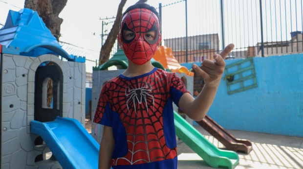 #ParaQueTodosPossamVer A imagem mostra um menino vestido de Homem-Aranha em um parquinho. Ele usa máscara e fantasia vermelha e azul, e faz o gesto clássico do herói com a mão, como se estivesse lançando uma teia. Ao fundo, há escorregadores coloridos e um muro pintado de azul. Foto: Prefeitura De Carapicuíba (Redes Sociais/Reprodução)